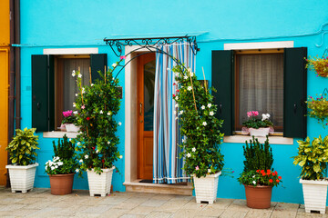 Fototapeta na wymiar Bright blue house on Burano island, Venice, Italy. Picturesque colorful wooden old style door and windows with green shutters and flowers on window sills
