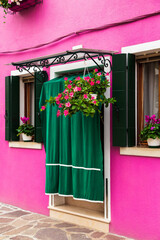 Fototapeta na wymiar Bright traditional pink house on Burano island, Venice, Italy. Green curtain on door, wooden old style windows with shutters and Mandevilla flowers on window sills