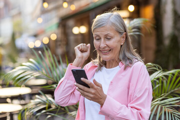 Senior woman outside, fist pumped in victory, smiling at her smartphone, capturing the joy of good...