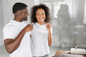 Happy African American millennial spouses brushing teeth together at bathroom