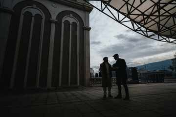 Two men engaged in a serious conversation outdoors by architectural columns.