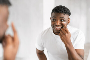 Cheerful millennial black guy applying moisturizer on face in bathroom