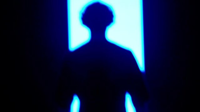 Male silhouette close up against digital wall in dark club. Man standing looking towards big digital screen background with neon strobing symbols, back shot.