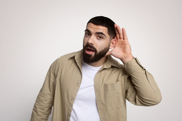 Obraz premium Curious young man with a beard, hand to ear, actively listening, expressing attention and interest
