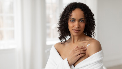 Beautiful black woman applying moisturising cream on shoulder in bathroom, lady wearing white bathrobe making beauty treatments at home, copy space