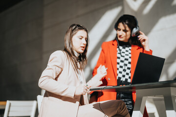 Two focused businesswomen engaging in a collaborative work session outdoors, using a laptop in a well-lit, urban setting.