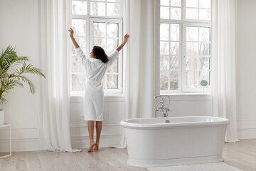 Young black woman in bathrobe opening curtains in light bathroom, lady standing near big window,...