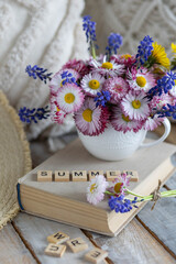 Colourful bouquet with blue flowers, meadow daisy in a cup on the table. Old book, straw hat, countryside home decor. Postcard for the holidays. Word summer written in wooden letters. Calendar