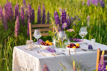 Romantic table decor for a loving couple on the blooming meadow with purple lupines. Two glasses of wine, flowers in a vase, silverware, fruits, wooden furniture and picnic basket. Sunset, golden hour