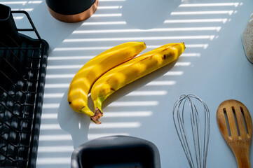 Banana on the kitchen counter illuminated in the shadow of the blinds. White natural light, friut background - 747594519