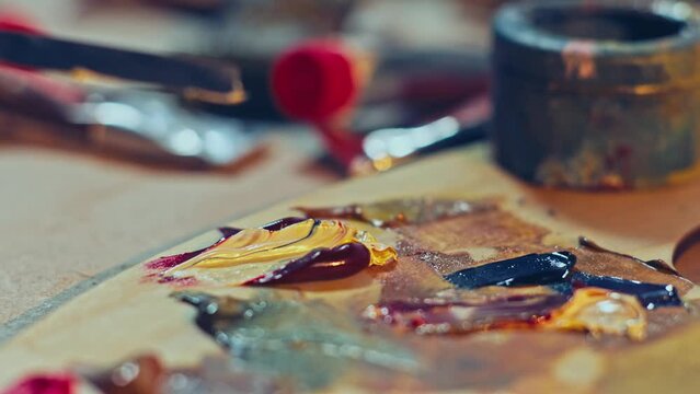 Artist mixing paints by a palette knife on the wooden palette. Red and yellow oil paints on the artist’s palette. Artist studio. 