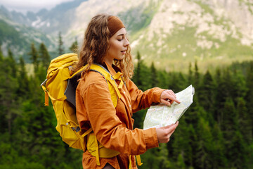 Stylish woman with backpack hiking, orient themselves to the terrain, study map and navigate the...
