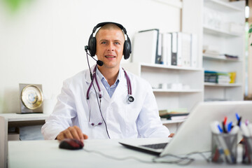 Portrait of positive confident doctor in headphones sitting at table in clinic office. Concept of...