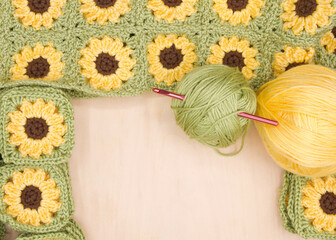Close up on piles of finished crochet granny squares, connected squares behind with balls of yellow and green yarn to side on light wood table. Crochet hook in green yarn ball. View from above