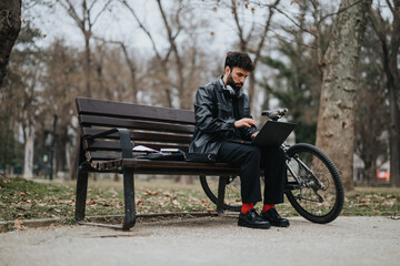 Focused male business entrepreneur using a laptop seated on a park bench with his bicycle beside him, embodying a blend of professionalism and casual outdoor work environment.