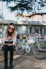 Naklejka premium Casual outdoor leisure concept with a trendy woman wearing sunglasses absorbed in reading while sitting on a swing, bikes parked behind.