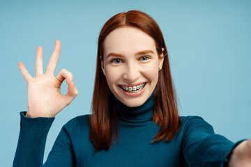 Smiling ginger woman with dental braces showing ok sign looking at camera
