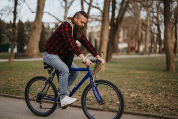 Casual bearded male riding a blue mountain bike on a park path surrounded by trees with autumn leaves