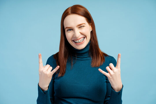 Funny overjoyed woman with braces wearing blue turtleneck showing rock sign looking at camera