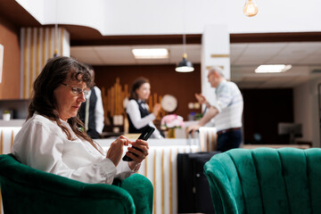 Fototapeta premium Caucasian senior female traveller relaxing on comfy sofa with smartphone in hotel lounge area. Retired senior woman looking for enjoyable holiday activities on her mobile device in beautiful resort.