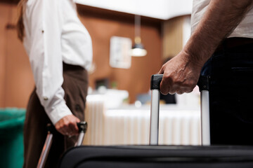 Close view of couple entering foyer of fancy hotel with trolley bags. Detailed shot of elderly individuals grasping suitcases as they walk up to registration desk to check in with the receptionists.
