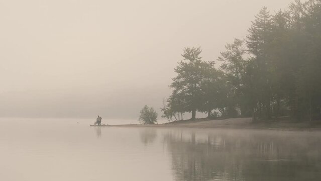 Silhouette of people fishing on the lake shore during foggy morning.