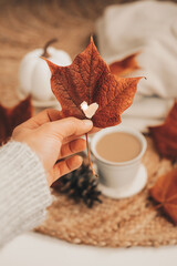 Hand holding autumn maple leaf with a heart shaped hole on a festive fall background with a cup of...