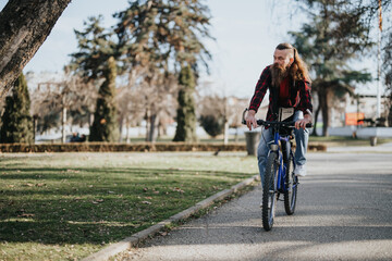 Bearded businessman in casual attire cycling in a city park with a laptop, showcasing a blend of work and eco-friendly transport.