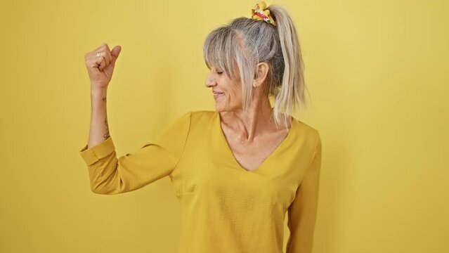 Strong middle age woman with grey hair in a pony tail flexing arm muscle on isolated yellow background, an epitome of power and fitness