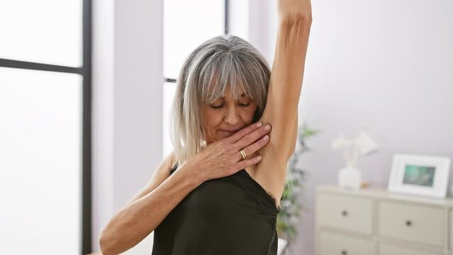 A mature woman with grey hair examines her underarm in the privacy of her modern bedroom.
