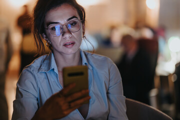 Lovely woman in glasses attentively checking her mobile phone in a low-light work environment,...