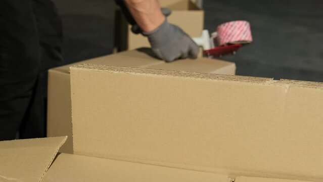 Worker uses parcel tape gun for packaging cardboard box with products. Man in gloves prepares box for distribution in storage