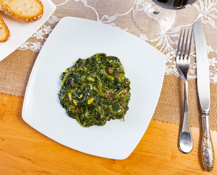 On table there is plate with stewed side dish, spinach in Catalan. Dish is steamed with addition of pine nuts and raisins. It is served with meat and fish dishes