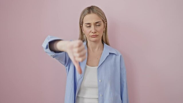 Angry young blonde in shirt displays negative thumbs down gesture, bad expression shows unhappy disgust. isolated over pink background