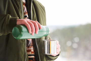 Unrecognizable male tourist pouring hot tea or water from vacuum flask into mug on cold winter day