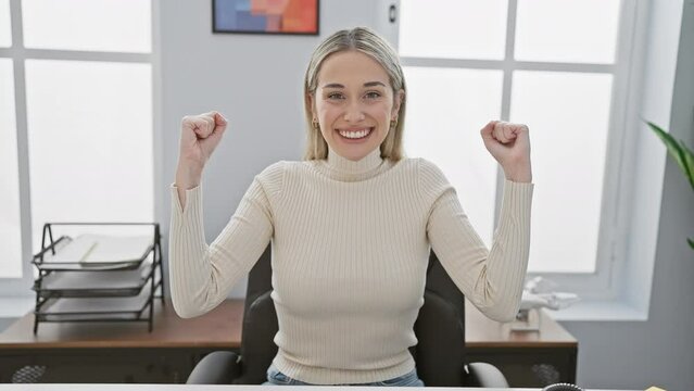 Joyful blonde woman celebrating win at office, triumphant expression of a young achiever sitting on table with a victorious, cheerful smile!