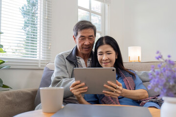 A senior couple in their 60s spends their free time sitting on a tablet, relaxing and watching...