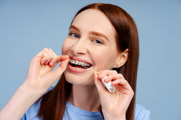 Ginger haired woman with dental brackets cleaning her teeth with floss on blue background
