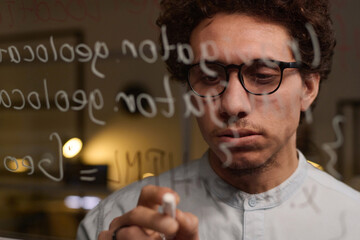 Closeup of young man wearing eyeglasses developing computer code writing it on glass wall
