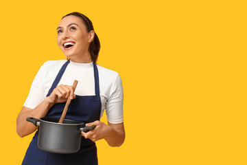 Portrait of happy young housewife in apron with cooking pot and wooden spoon on yellow background