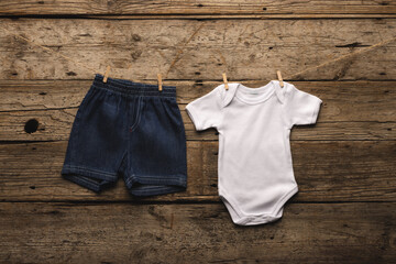 A pair of baby denim shorts and a white onesie are pinned to a clothesline against a rustic wooden b