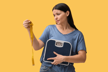 Beautiful young displeased woman with measuring tape and scales on yellow background. Weight loss...