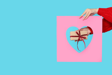 Female hands holding gift box and paper sheet with hole in shape of heart on blue background....