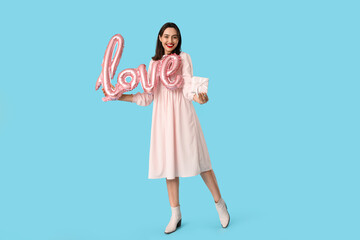 Beautiful young happy woman with gift box and balloon in shape of word LOVE on blue background. Valentine's Day celebration
