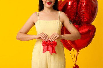 Beautiful young happy woman with gift box and heart-shaped balloons on yellow background....