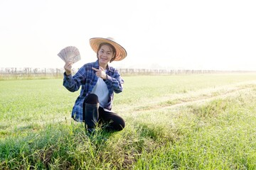 A female Asian farmer sitting and point to the amount of Thai banknotes in the middle of a rice field at dawn.