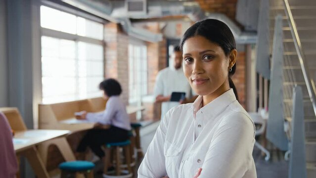 Portrait of young businesswoman with serious expression working in modern open plan office turning to look at camera and folding arms with colleagues in background - shot in slow motion