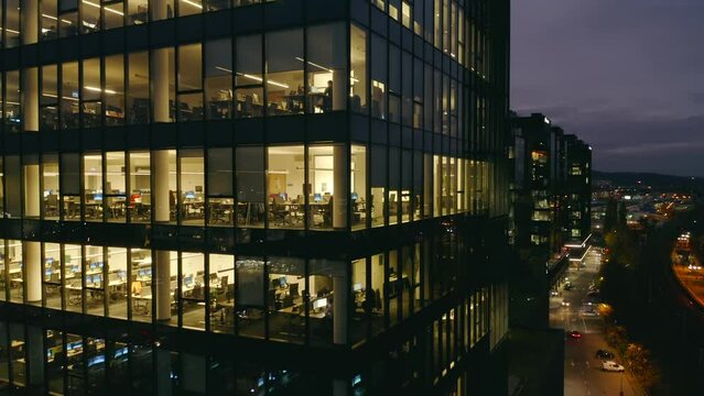 Modern office building at night. Aerial view of the windows of a multi-story skyscraper building made of glass, office lighting and people working until late in the evening inside.