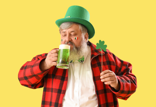 Mature man in hat with paper clover drinking beer on yellow background. St. Patrick's Day celebration