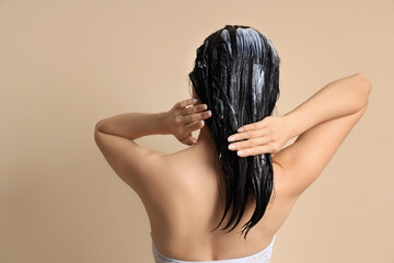 Young woman applying hair mask on beige background, back view